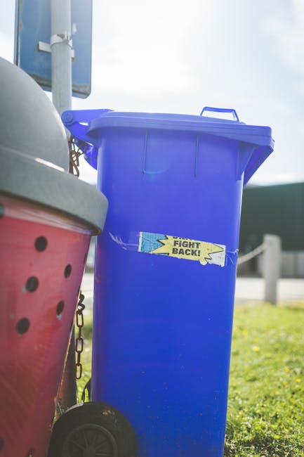 A tall, bright blue plastic rubbish bin with a hinged lid is positioned outdoors on a pavement or driveway, with a small yellow sticker on the front that reads 'FIGHT BACK!'. The bin's surface appears smooth and glossy, reflecting natural daylight, and it is placed adjacent to a red container with black ventilation holes, which is partly visible on the left side of the image. The ground nearby is grassy, and there are metal barriers or posts in the background, suggesting an urban or suburban environment. The sky overhead is partly cloudy, with sunlight illuminating the scene, emphasizing the vibrant color of the blue bin. This scene relates to private waste collection services, where alternative rubbish disposal solutions are employed outside of local authority collection, highlighting the role of independent waste management providers such as Waste Collection Stockwell in managing household or commercial waste in the area.