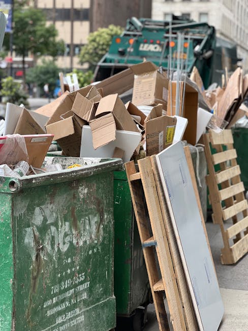 A large green waste disposal dumpster filled with various discarded cardboard boxes, some flattened and others partially assembled, alongside wooden pallets leaning against its side. The boxes are made of brown and white cardboard, with some showing printed markings or labels. The pallets are light-colored, with slatted wooden planks arranged in a rectangular frame, positioned vertically next to the dumpster. The scene is set outdoors on a paved urban street, with a municipal waste collection vehicle visible in the background, partially obscured by the pile of rubbish. Surrounding the scene are trees, buildings, and parked cars, indicating a city environment. The lighting appears natural, with daylight providing even illumination across the scene, emphasizing the textures of the materials and the compacted nature of the waste. This image illustrates typical arrangements for private waste disposal or rubbish removal services, as provided by companies like Waste Collection Stockwell, highlighting the process of on-site clearance and collection of assorted packaging and reusable pallet materials.