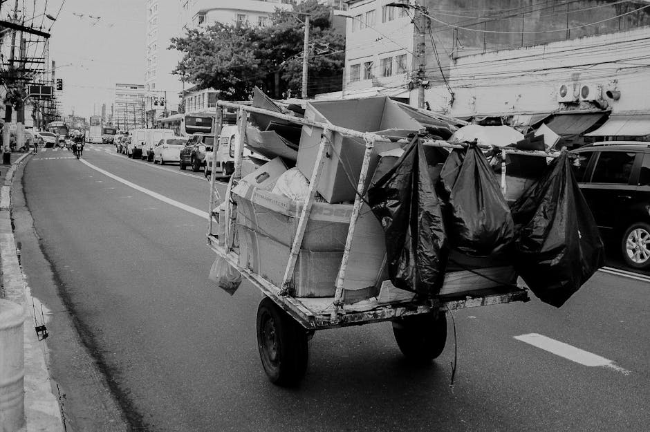 A mobile load of discarded household items and rubbish carts on a city street, featuring a small, flatbed trolley filled with various bundled materials. The trolley is constructed with a metal frame and has two large rear wheels, supporting an assortment of large cardboard boxes, plastic bags, and smaller debris. Some of the black plastic rubbish bags are strapped or tied to the sides of the trolley, hanging loosely. The background shows vehicles parked along the curb, including sedans and SUVs, with a row of multi-storey buildings displaying residential or commercial windows and external air conditioning units. The street surface is paved with asphalt, marked with white lane lines, and the environment is lit with natural daylight, creating a clear, neutral scene. The image depicts an informal or alternative approach to rubbish collection, which Waste Collection Stockwell might facilitate through private waste handling or on-site clearance services for cluttered urban environments.