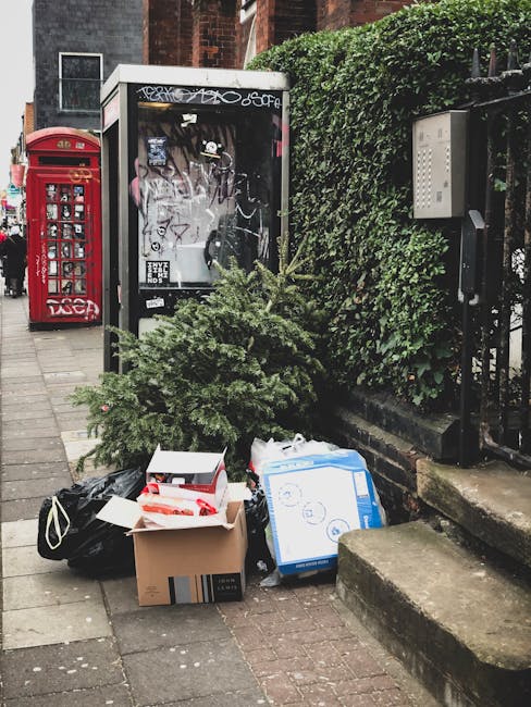 A pile of waste materials including a small, green, artificial Christmas tree, cardboard boxes, plastic bags, and a white boxed item is arranged on a paved sidewalk in front of a black iron fence with an overgrown green hedge. Behind the waste, there is a glass-fronted telephone booth with graffiti at the top, alongside a traditional red British post box. The scene is set in an urban area with brick building facades visible in the background, indicating a typical street scene where private rubbish disposal, managed by Waste Collection Stockwell, might be necessary for alternative waste handling or on-site clearance.