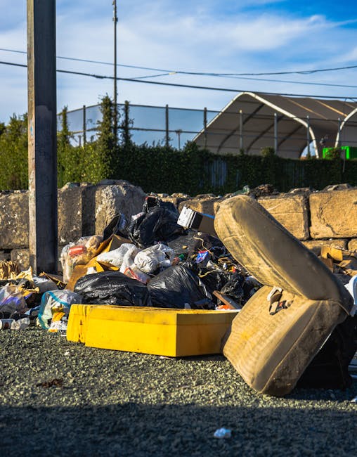 A mobile load of discarded household items and rubbish carts on a city street, featuring a small, flatbed trolley filled with various bundled materials. The trolley is constructed with a metal frame and has two large rear wheels, supporting an assortment of large cardboard boxes, plastic bags, and smaller debris. Some of the black plastic rubbish bags are strapped or tied to the sides of the trolley, hanging loosely. The background shows vehicles parked along the curb, including sedans and SUVs, with a row of multi-storey buildings displaying residential or commercial windows and external air conditioning units. The street surface is paved with asphalt, marked with white lane lines, and the environment is lit with natural daylight, creating a clear, neutral scene. The image depicts an informal or alternative approach to rubbish collection, which Waste Collection Stockwell might facilitate through private waste handling or on-site clearance services for cluttered urban environments.
